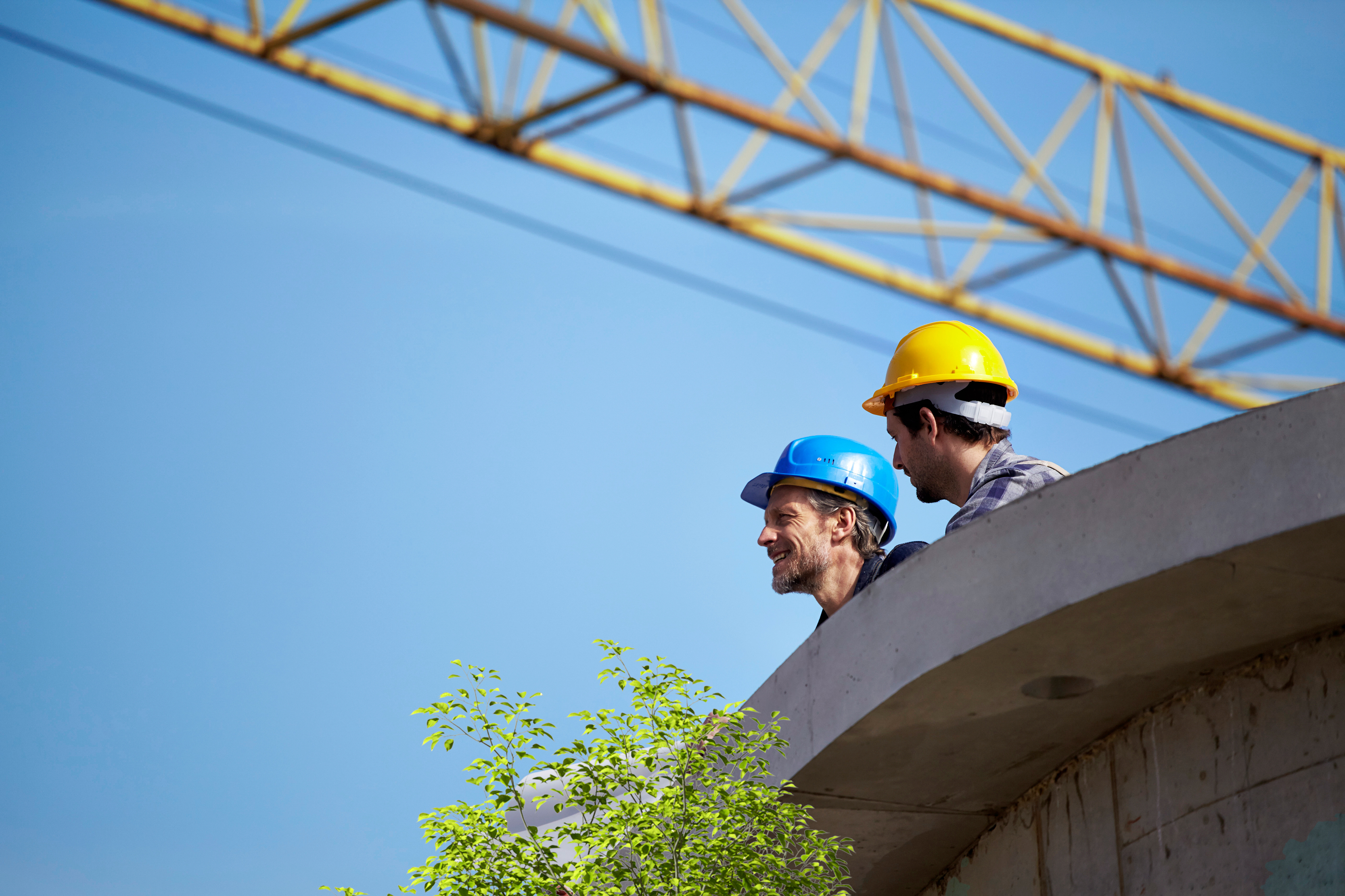 Zwei Bauarbeiter blicken mit einem Kran im Hintergrund von einer Baustelle 