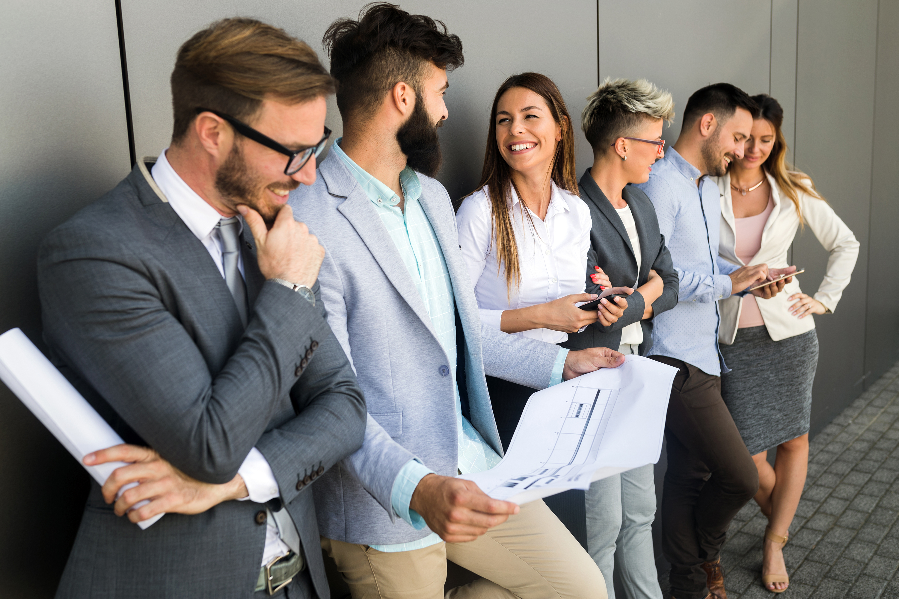 Ein großes Team von Männern und Frauen unterhält sich lachend an eine Bürowand gelehnt